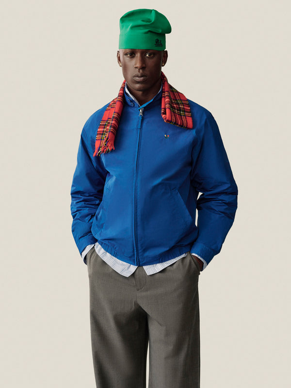 Man in blue jacket, red plaid scarf, Unisphere Skull Cap, gray pants stands against a plain background.