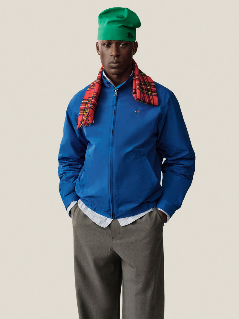 A man in a blue jacket and green hat wears Houndstooth Elasticated Waist Trousers, hands in pockets, plain background.