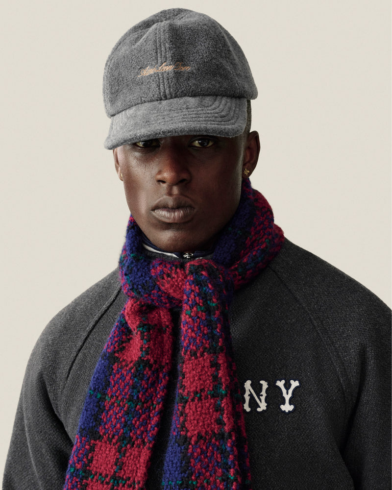 A person wearing a grey fleece Script Fleece Hat with gold embroidery and a red and blue plaid scarf.