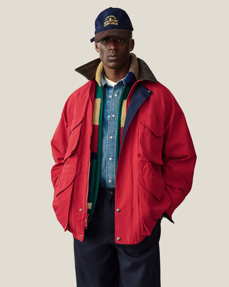 Man in Lightweight Barn Jacket, blue denim shirt, striped scarf, navy pants, and navy cap stands by a plain backdrop.