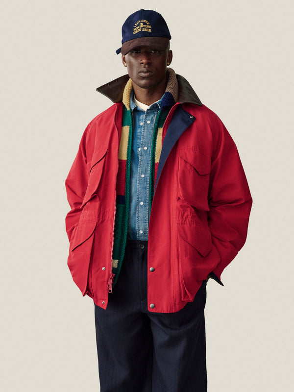 Man in red jacket, denim shirt, striped scarf, navy pants, and Triborough Chess Club hat stands against plain background.