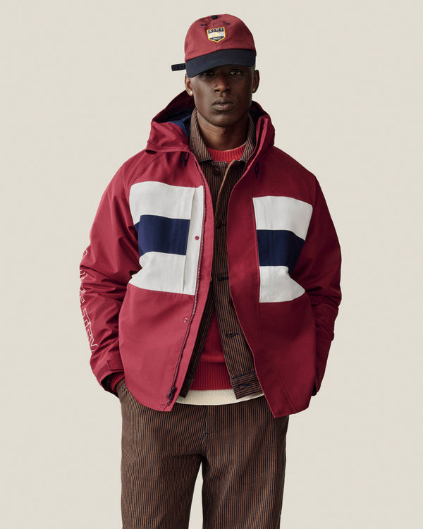 A man wears a GORE-TEX Striped Sailing Jacket with white/navy panels, a matching cap, red sweater, and brown pinstripes.