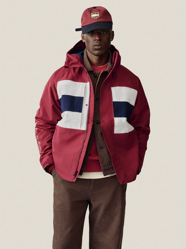 Man in a red hooded jacket, brown striped pants, and Crest Shield Hat stands facing forward.