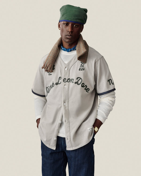 Man in a Mulberry Chainstitch Baseball Jersey, green beret, plaid scarf, white shirt, and blue striped pants.