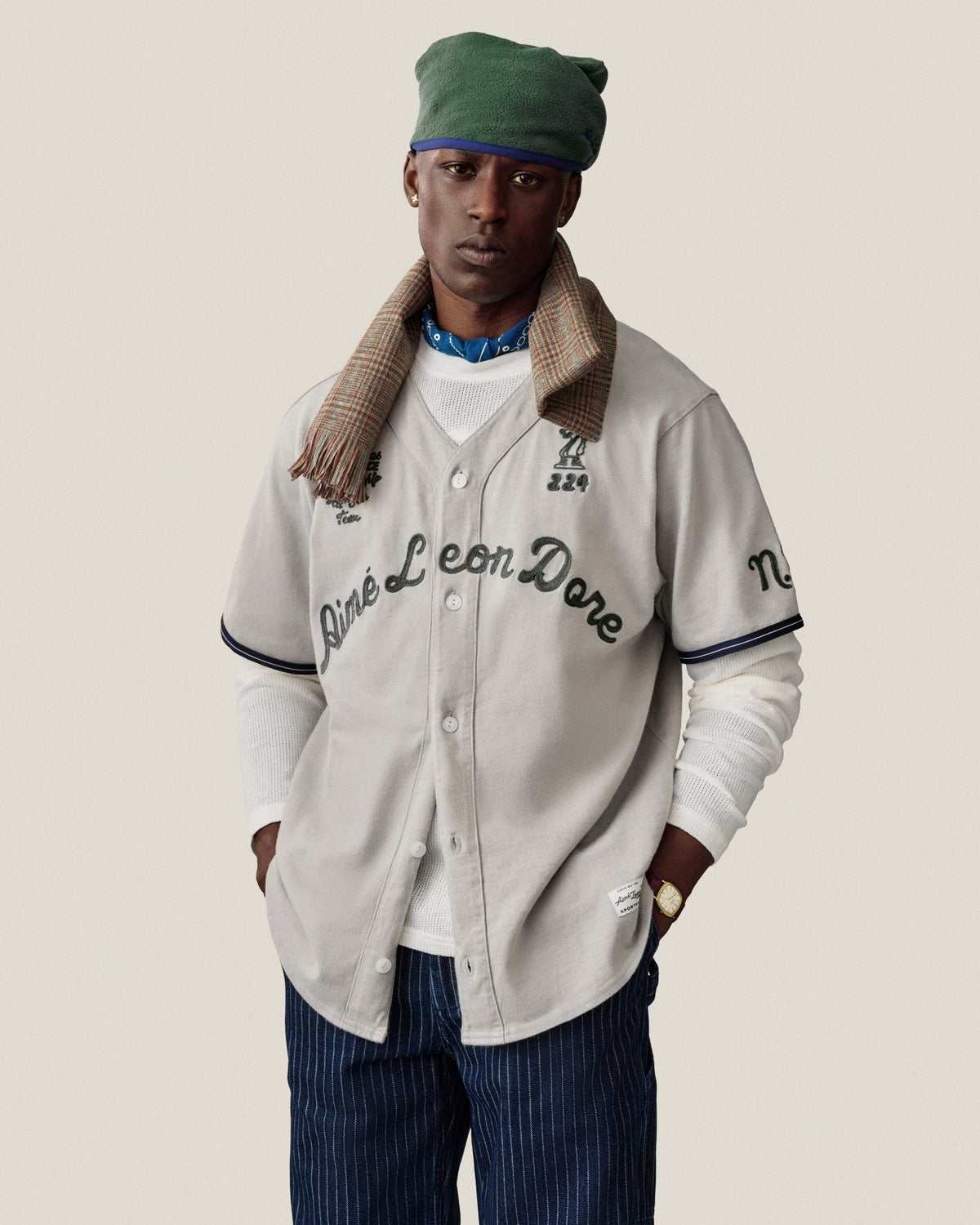 A man wears a cream Aimé Leon Dore baseball jersey, green cap, plaid scarf, white tee, and Chalk Stripe Painter Pant.