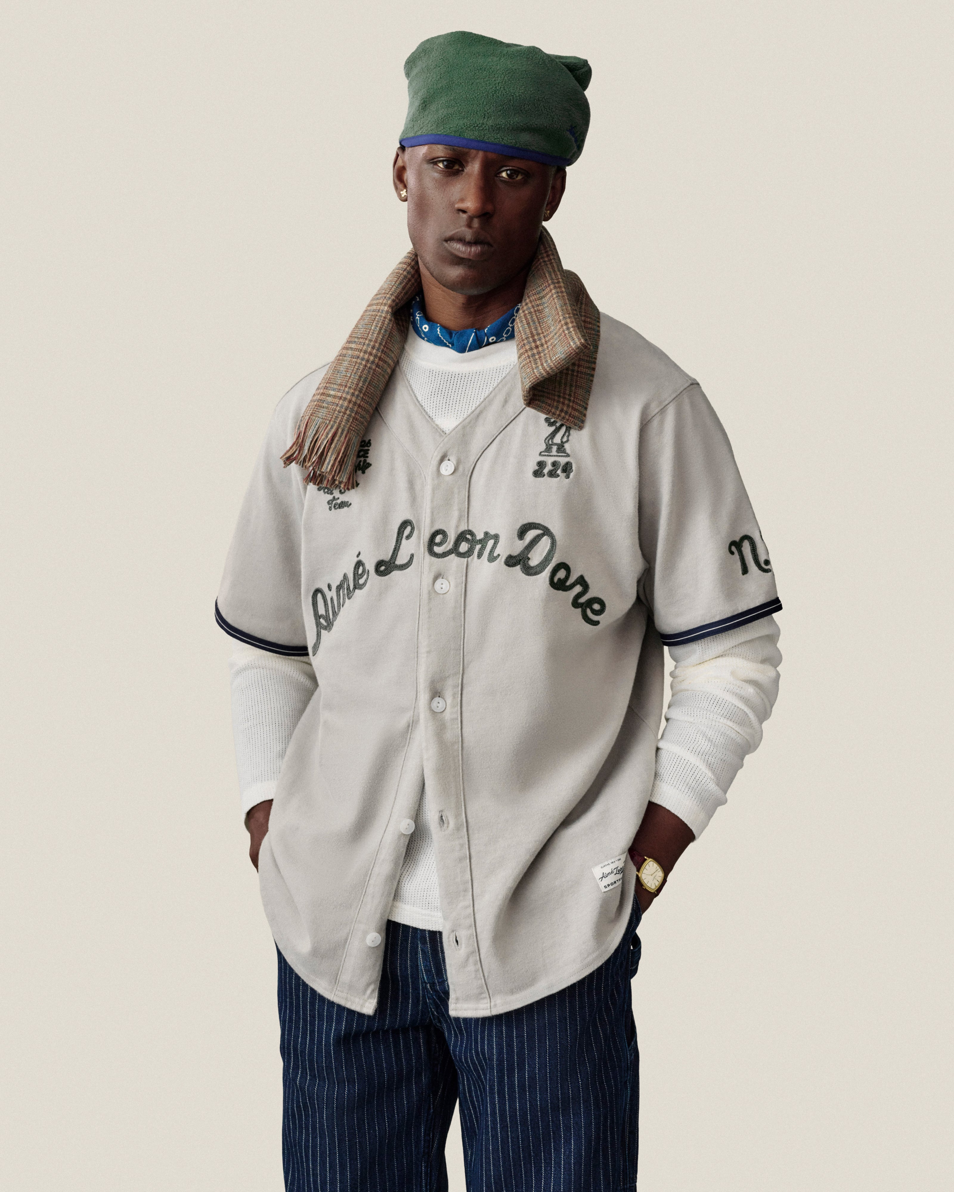 A man wears a cream Aimé Leon Dore baseball jersey, green cap, plaid scarf, white tee, and Chalk Stripe Painter Pant.