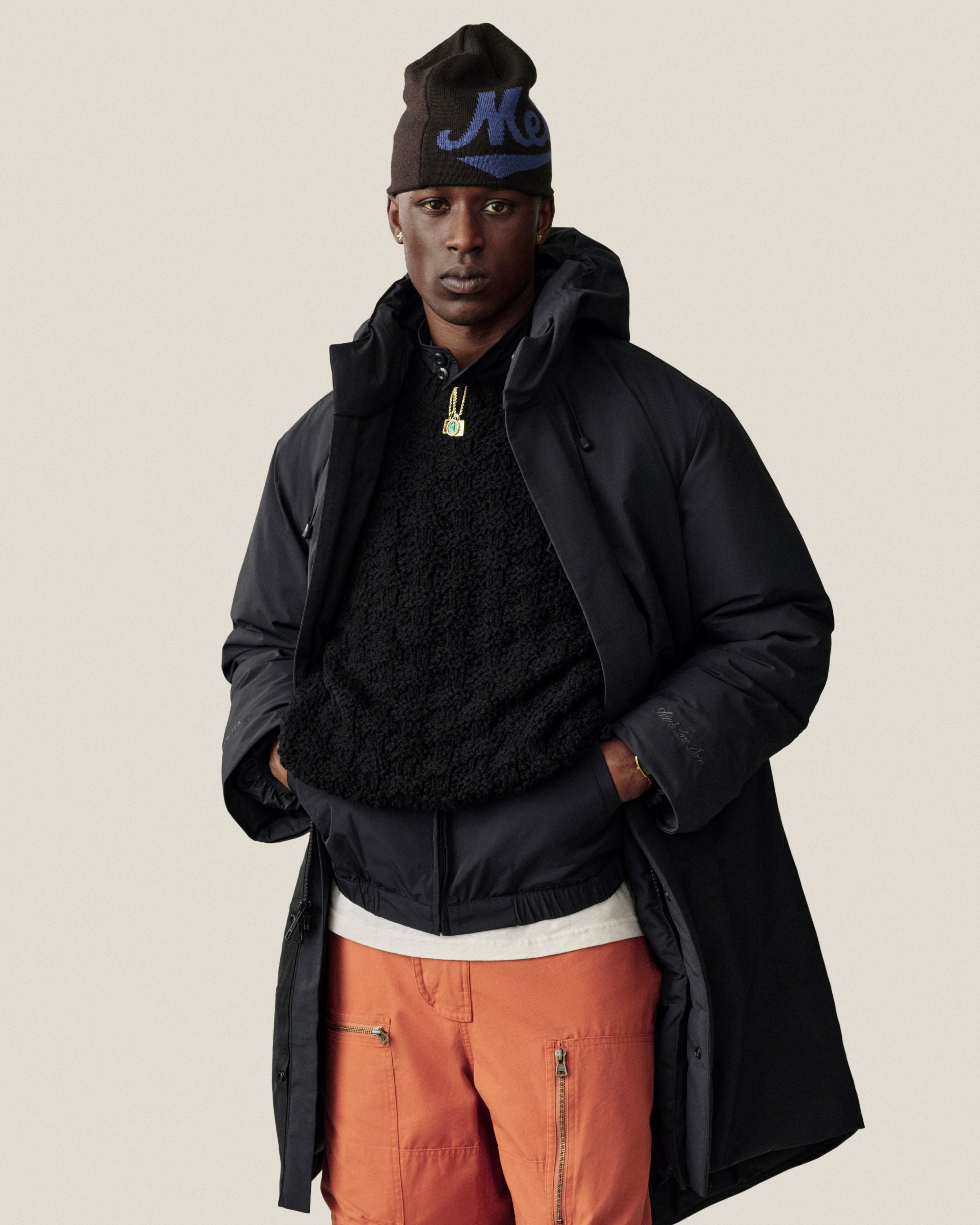 A person wears an ALD / New Era Mets Beanie, a black hooded coat, a black fleece, and orange pants.