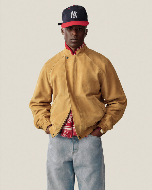 Man in a suede cafe racer jacket, red scarf, blue jeans, and Yankees cap against a plain background.