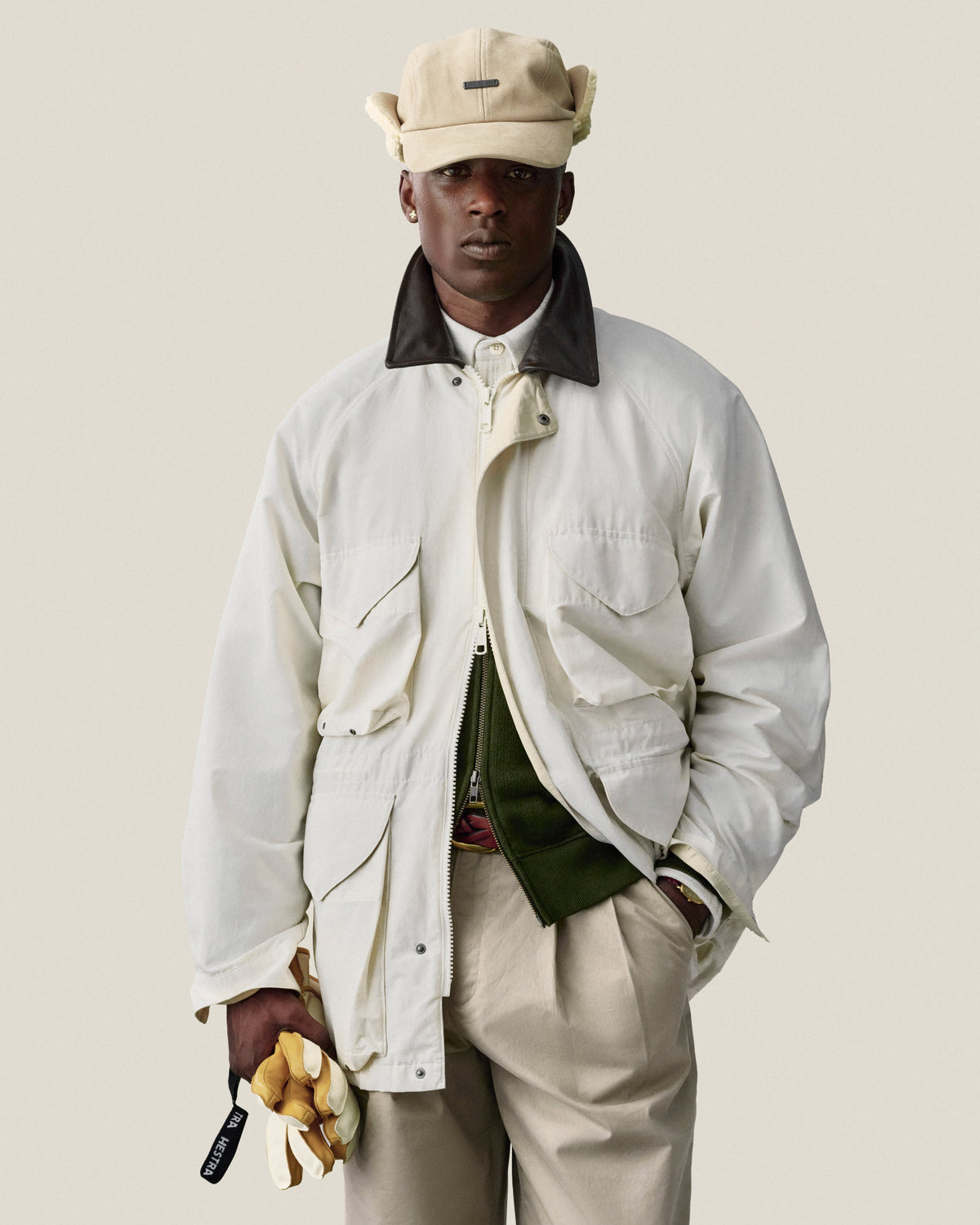 Man in Double Pleated Officer Pant, cream jacket, and hat stands against a plain light background, holding gloves.