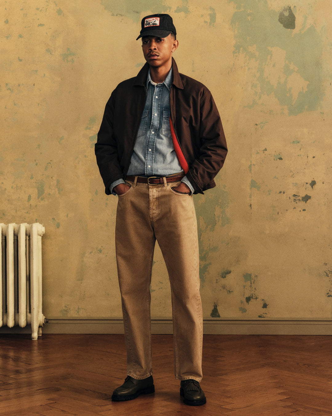 A man wearing an Aimé Retriever Hat, a dark brown jacket over a denim shirt, and tan pants.