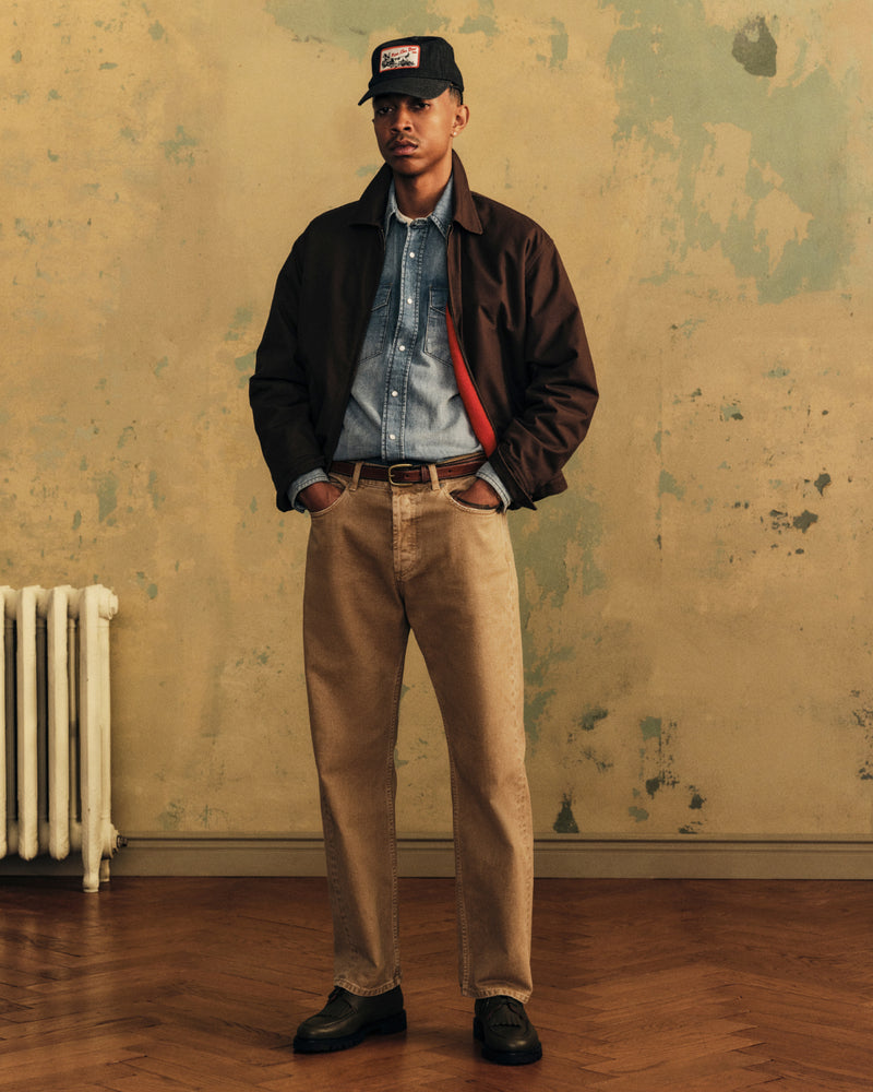 A man wearing an Aimé Retriever Hat, a dark brown jacket over a denim shirt, and tan pants.
