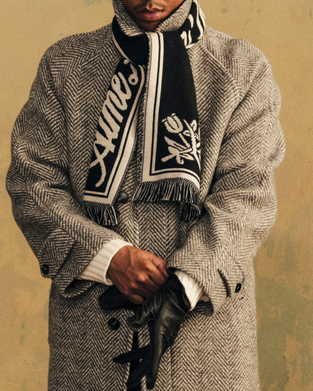 Wool Script Scarf in black with white text and a floral design, worn over a gray herringbone coat.