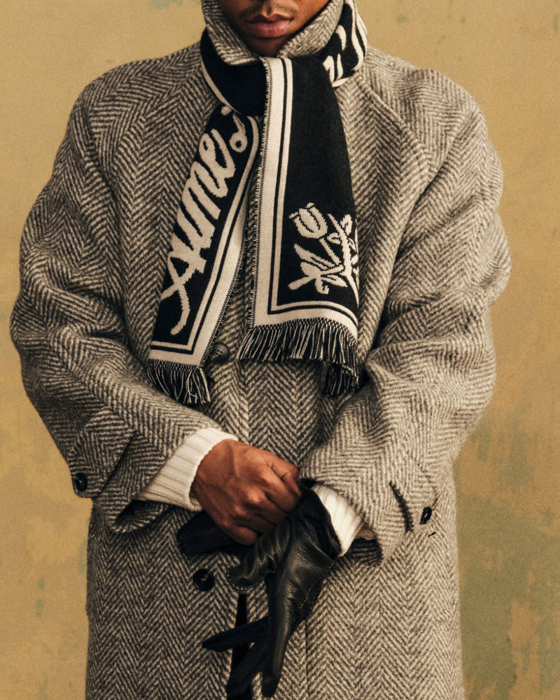 Wool Script Scarf in black with white text and a floral design, worn over a gray herringbone coat.