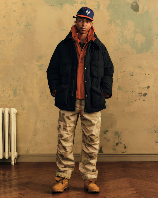 Person wearing a black Down-Filled Surplus Parka over an orange hoodie, camouflage pants, and a baseball cap.