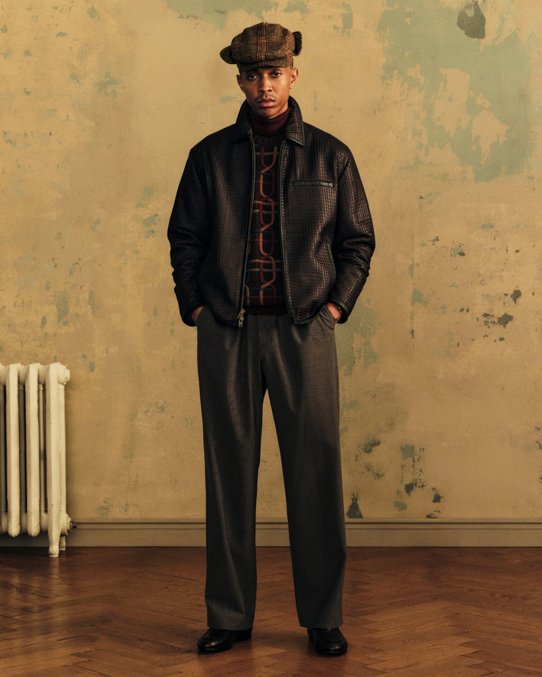 A man wearing a Plaid Tweed Driver’s Hat, a dark patterned jacket, and gray trousers stands in a room.