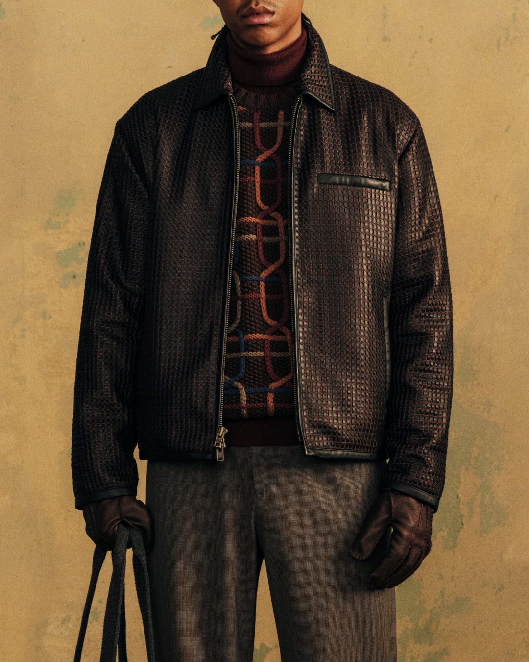 A person wears a textured dark jacket over a multi-colored Cable Link Crewneck Sweater and grey trousers.