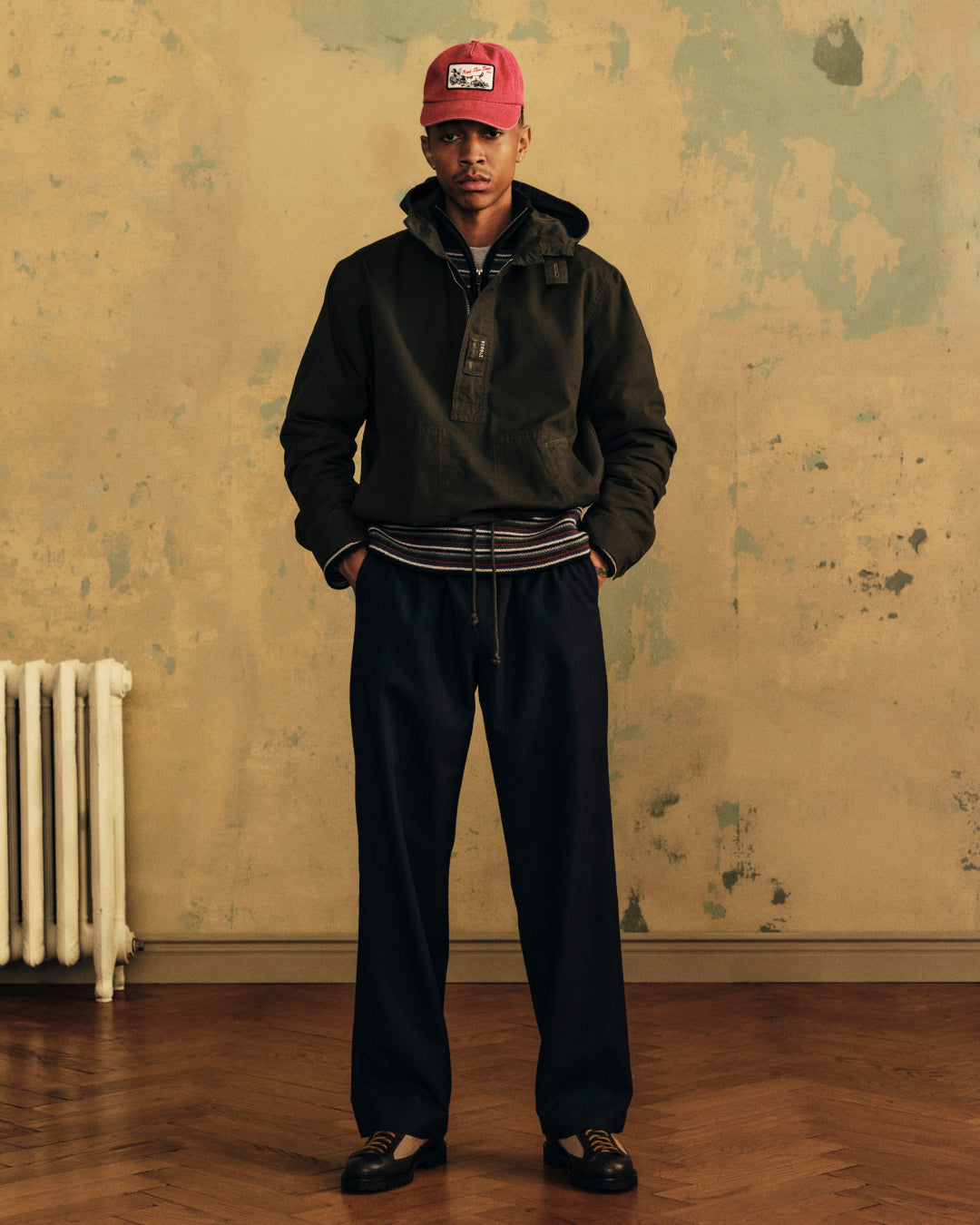 A person wearing a dark Garment Washed Anorak, red baseball cap, and wide-leg trousers stands with hands in their pockets.