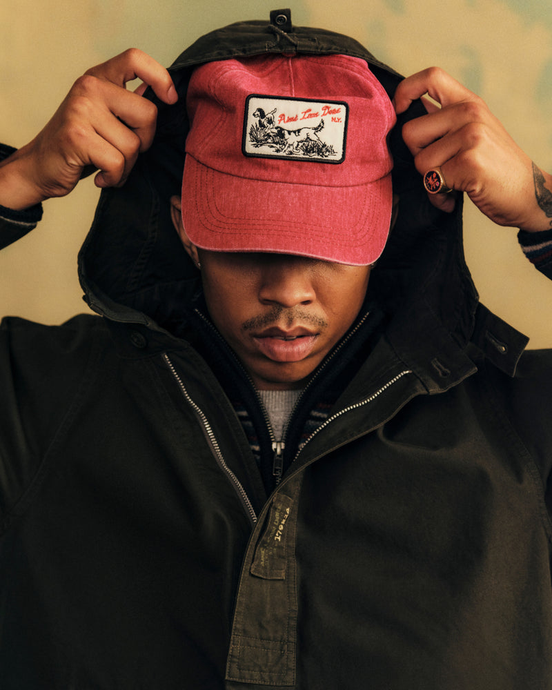 A person wearing a red Aimé Retriever Hat with a dog patch, pulling up a dark jacket hood.