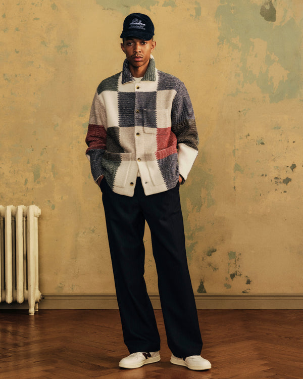 A person wearing a multicolored Patchwork Knit Chore Jacket, dark trousers, a baseball cap, and sneakers.