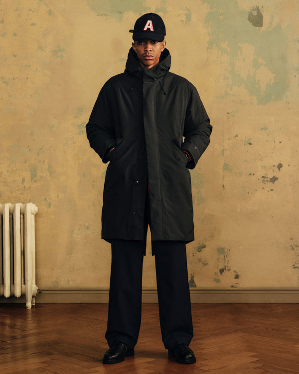 A person wearing a black Wool Varsity Logo Hat, a dark hooded parka, and black trousers.