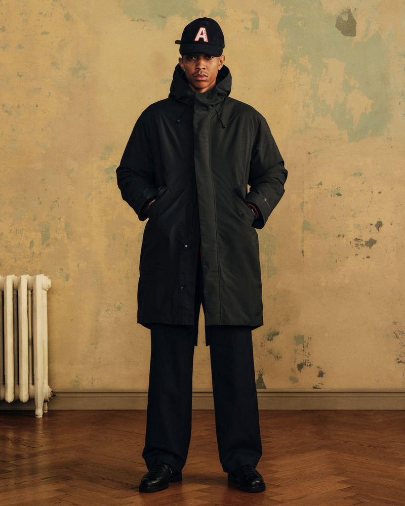 A person wearing a black Wool Varsity Logo Hat, a dark hooded parka, and black trousers.