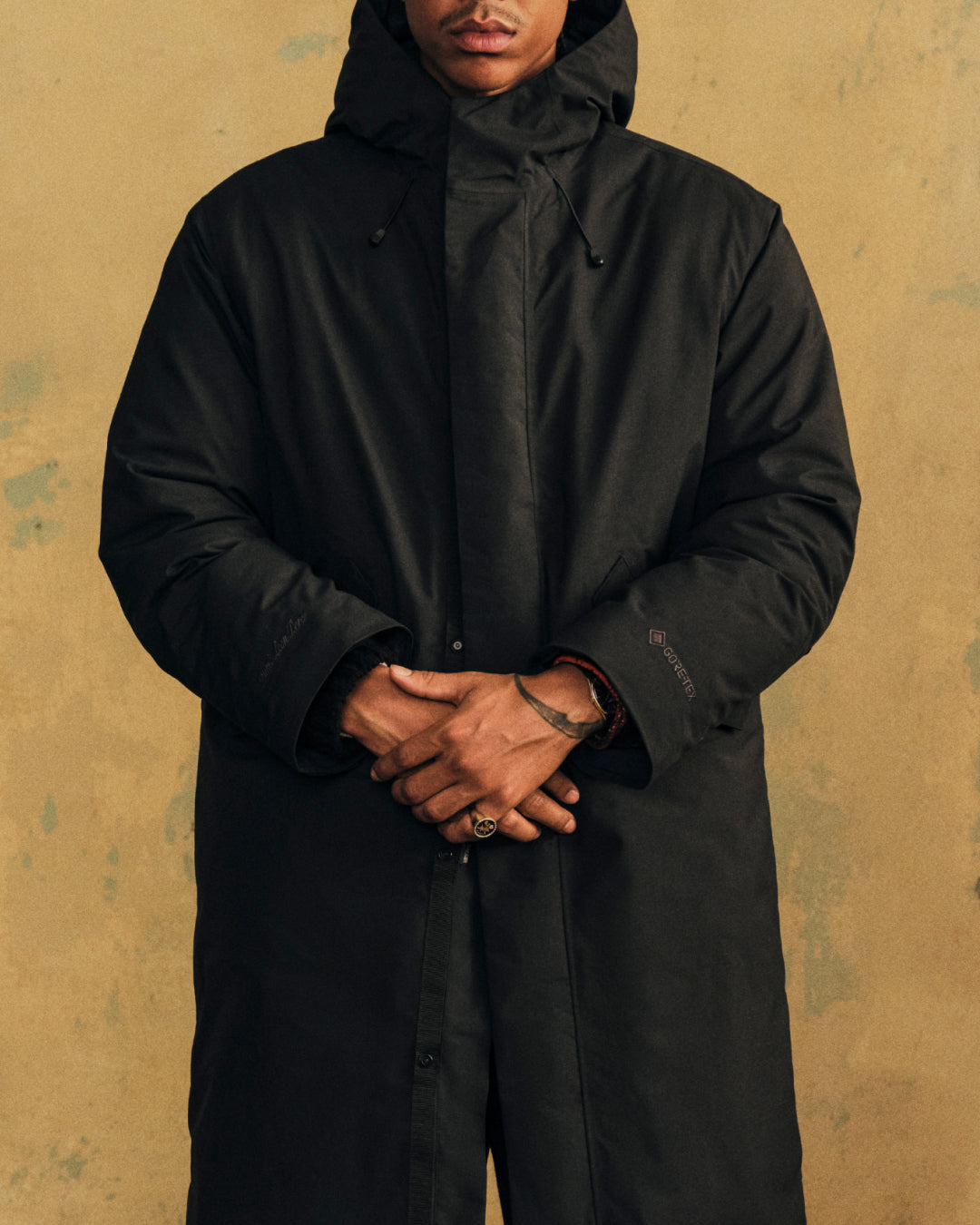 A person wearing a black GORE-TEX Down Filled Fishtail Parka with a hooded, down-filled design and a fishtail hem.