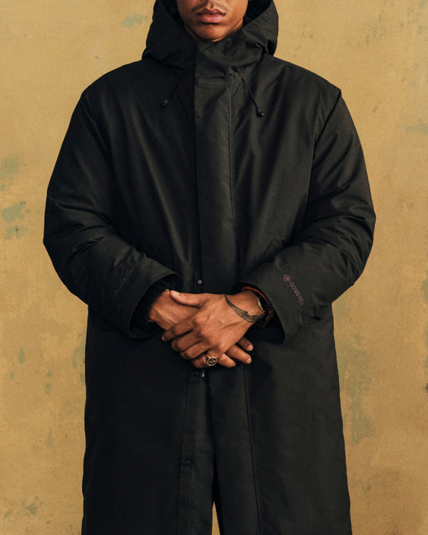 A person wearing a black GORE-TEX Down Filled Fishtail Parka with a hooded, down-filled design and a fishtail hem.