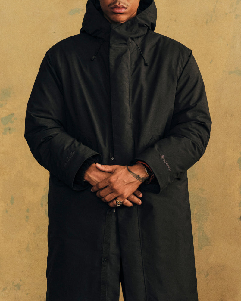 A person wearing a black GORE-TEX Down Filled Fishtail Parka with a hooded, down-filled design and a fishtail hem.