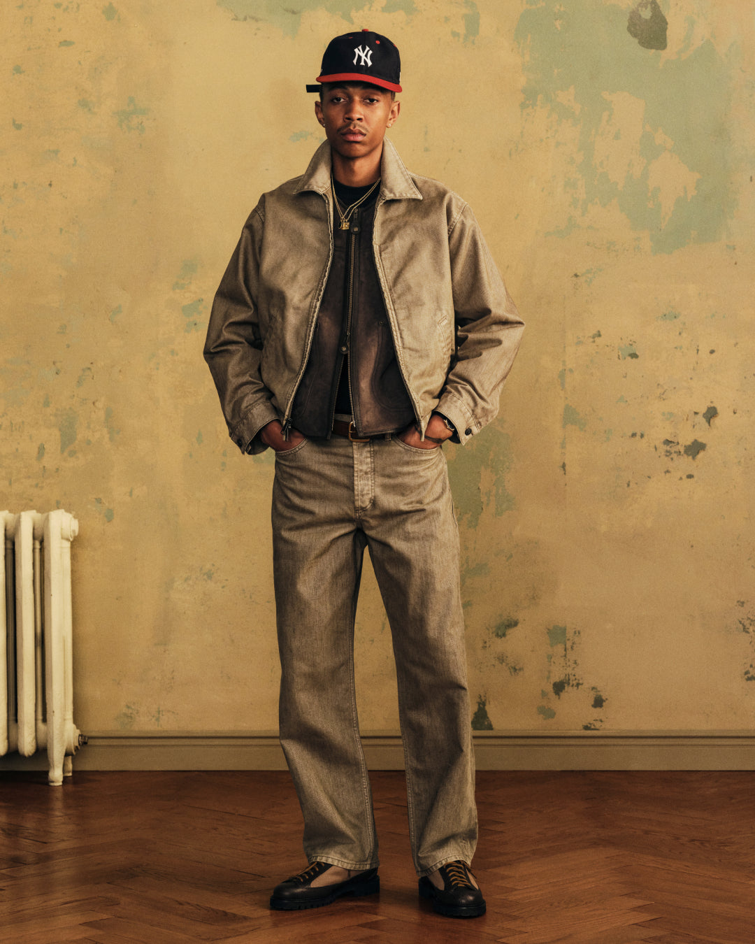 A man wears a tan jacket and Flocked Straight Fit Denim with a baseball cap and dark shoes, hands in his pockets.