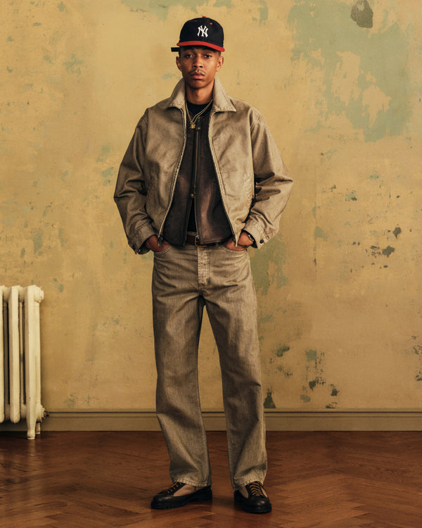 A man wears a tan jacket and Flocked Straight Fit Denim with a baseball cap and dark shoes, hands in his pockets.