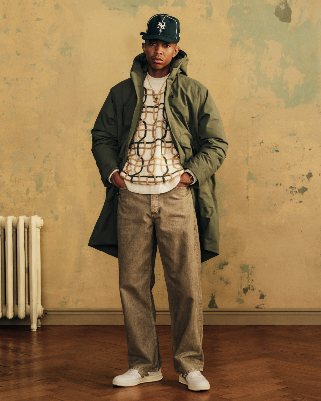 A man in a green parka, patterned sweater, and ALD / New Balance T500 Tennis Oxfords stands against a weathered wall.