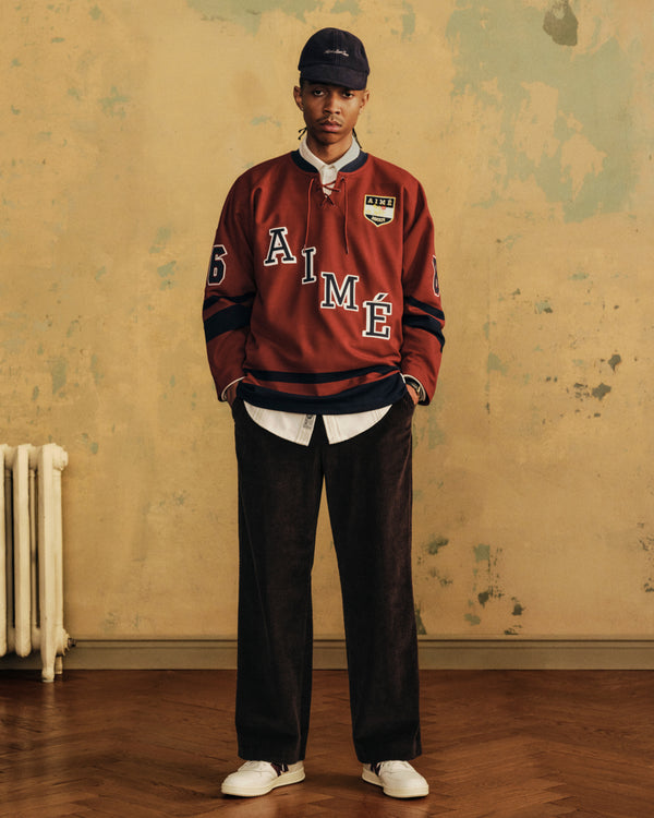 A person in a red lace-up jersey, collared shirt, and dark trousers wearing ALD / New Balance T500 Tennis Oxfords.