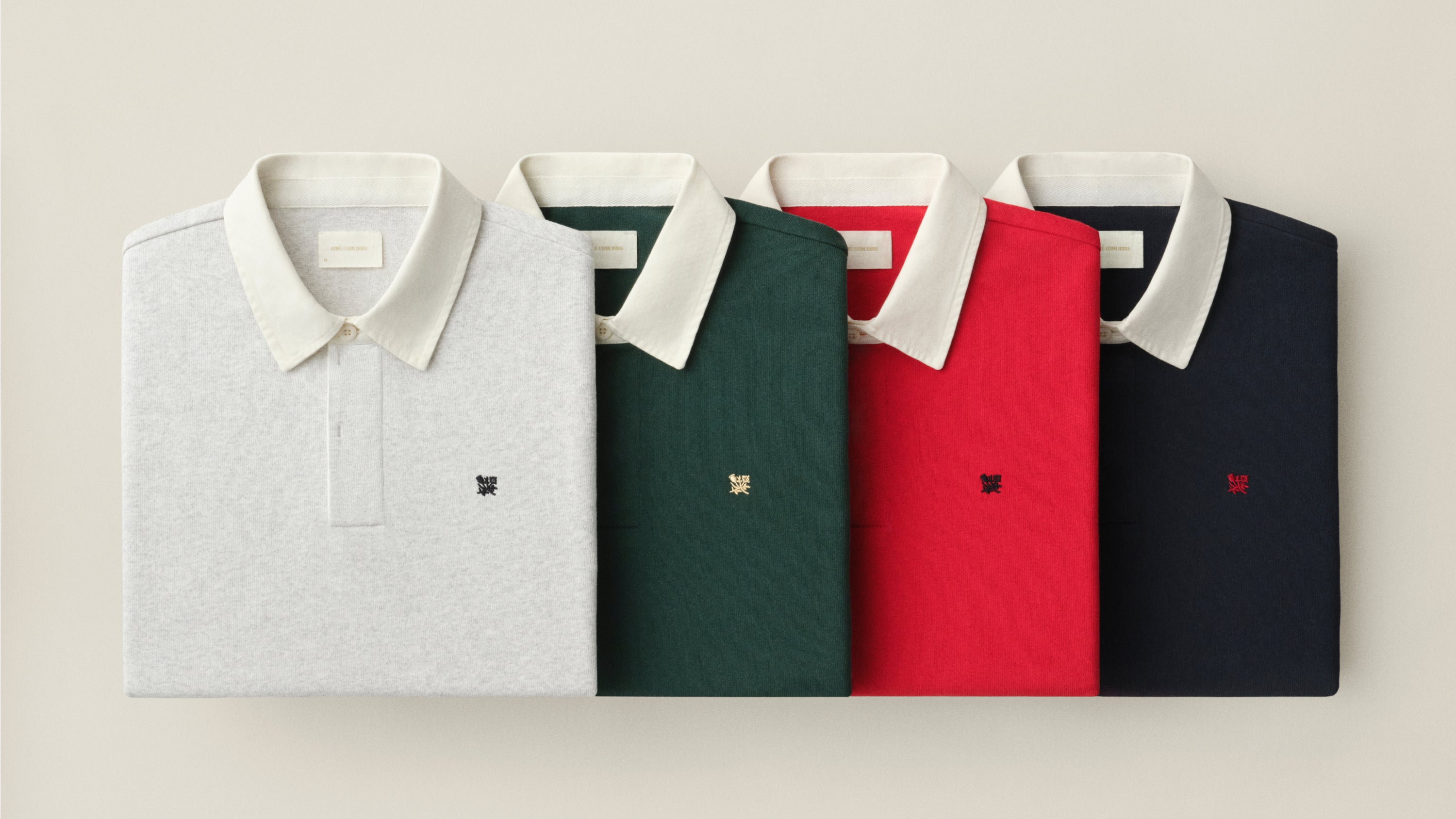 Four folded polo shirts with white collars are arranged side by side on a light beige background. From left to right, the shirt colors are light gray, dark green, bright red, and navy blue.