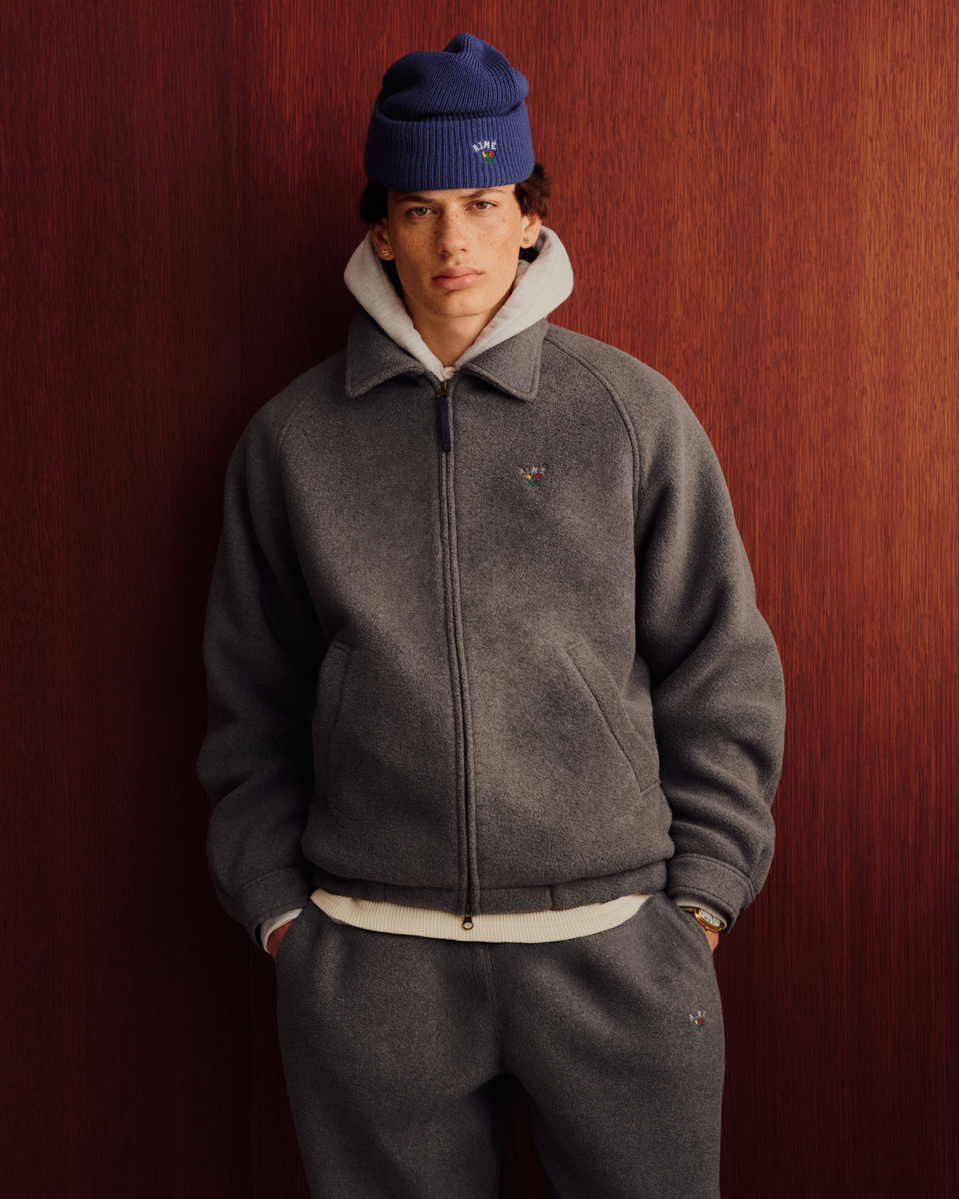 Someone in Crest Fleece Sweatpants, a gray zip jacket, and a blue beanie stands by a wood-paneled wall.