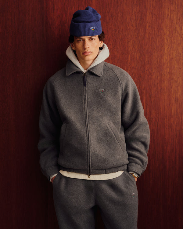 Man in gray jacket, sweatpants, Crest Fleece Full-Zip Hoodie, and blue beanie stands by a wooden wall, hands in pockets.