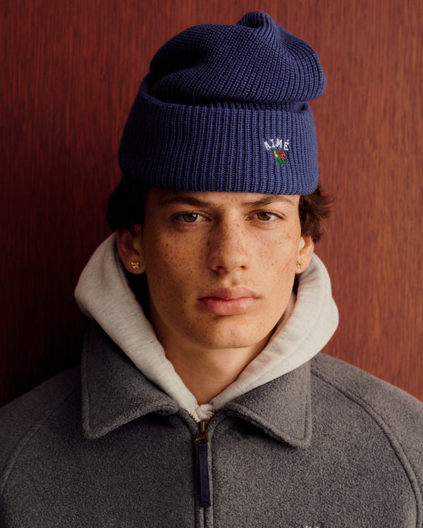 Young person in Crest Merino Wool Beanie, gray zip-up jacket, and hoodie stands by a wooden background.
