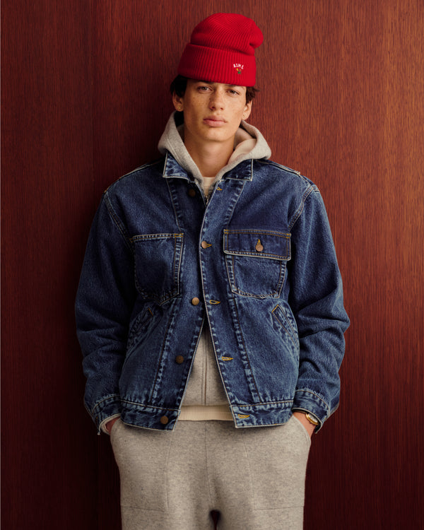 Young man in a red beanie and denim jacket wears a Full-Zip Pavilion Cashmere Hoodie, standing by a wooden wall.