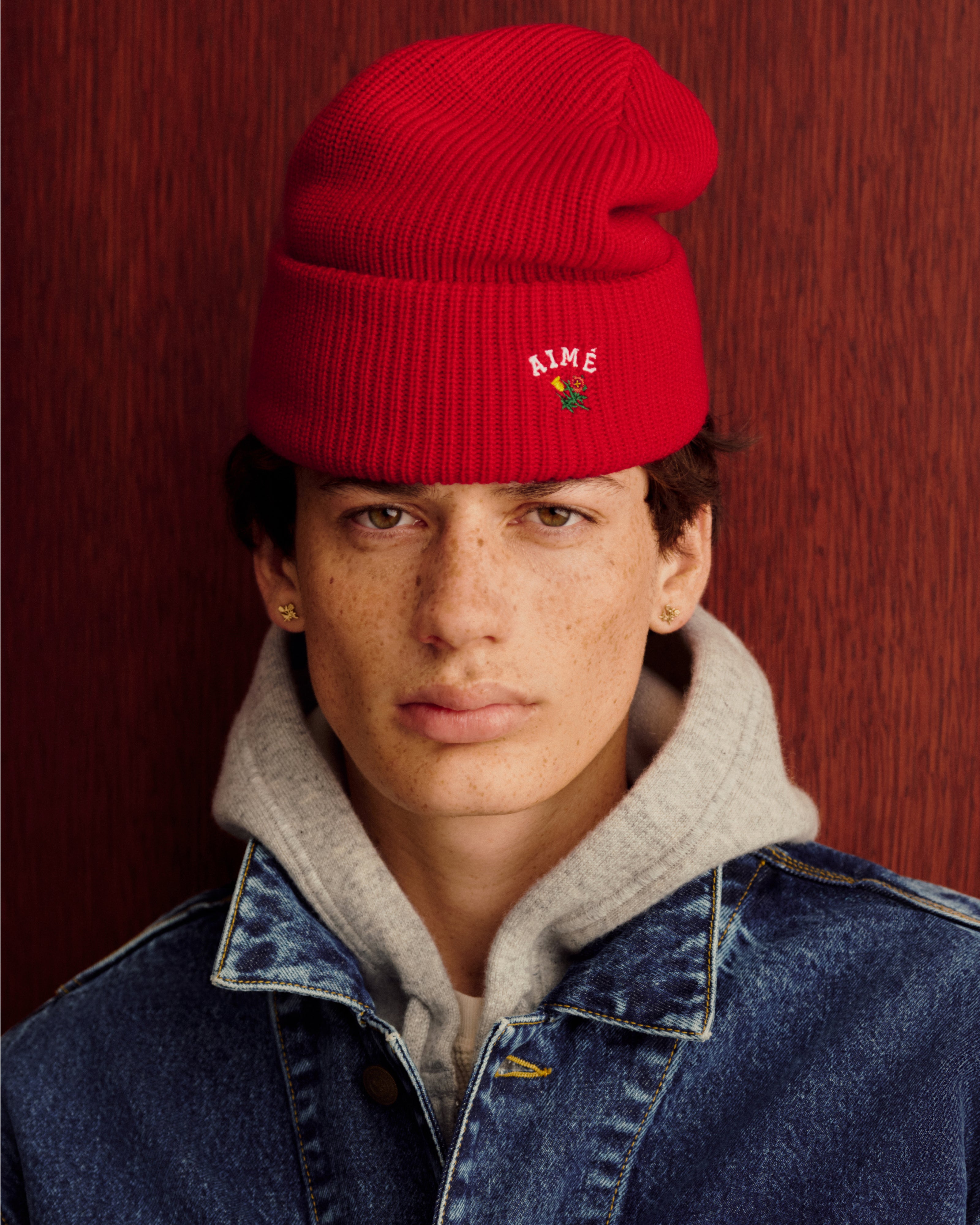 Someone in a Crest Merino Wool Beanie, gray hoodie, and denim jacket stands by a wood-textured background.