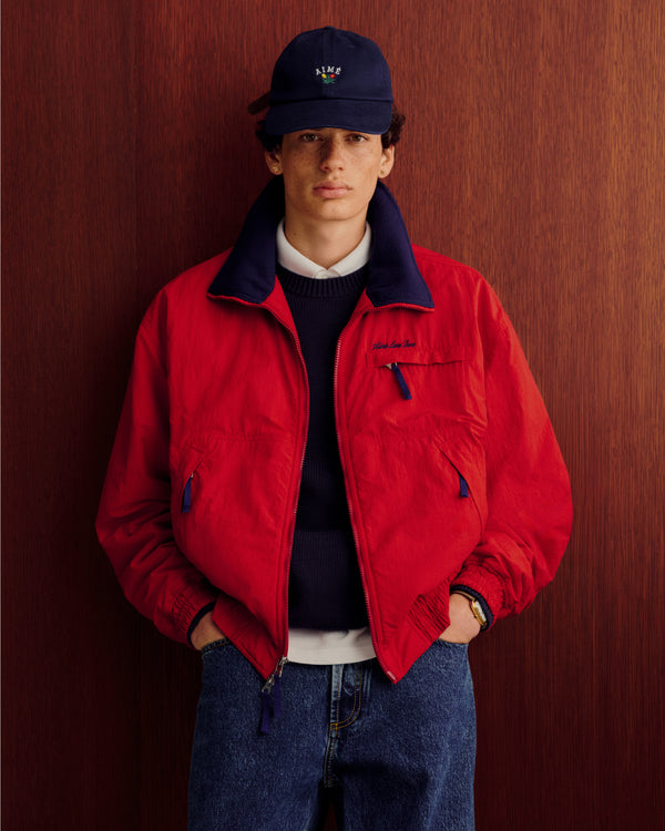 A person in a Crest Raglan Sweater, navy cap, and jeans stands before a wood-paneled background.