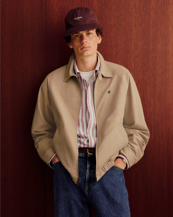 Young man in Striped Crest Oxford Shirt, beige jacket, jeans, and maroon cap stands by a wooden wall, hands in pockets.