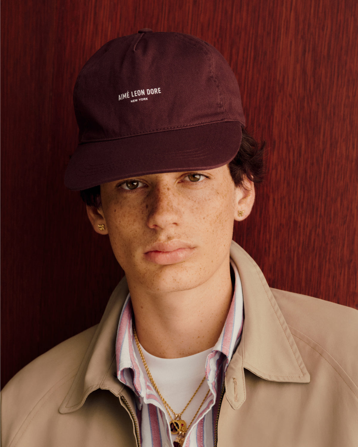Young person in a brown Micro Logo Hat, beige jacket, and striped shirt against a reddish wood background.
