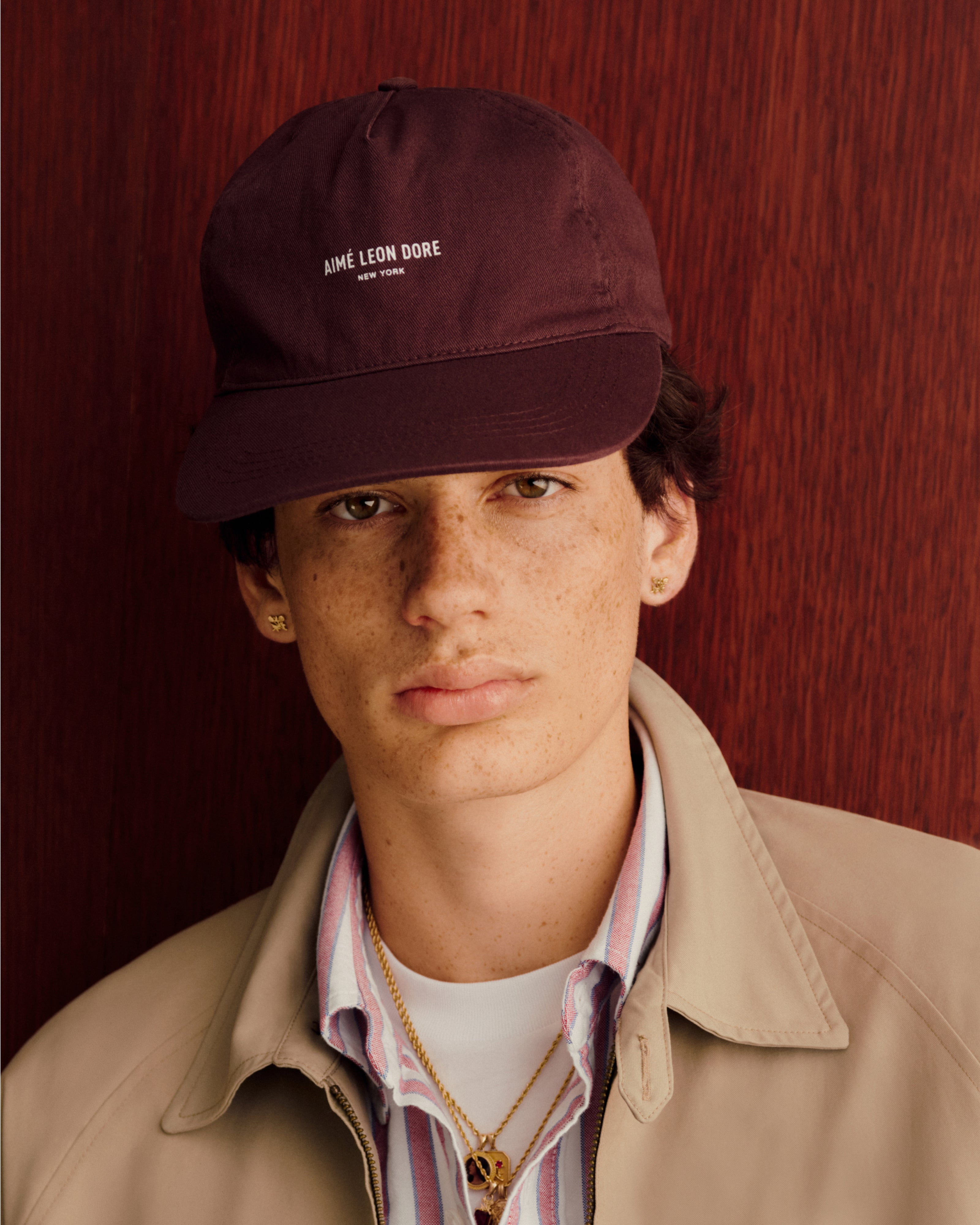 Young person in a brown Micro Logo Hat, beige jacket, and striped shirt against a reddish wood background.