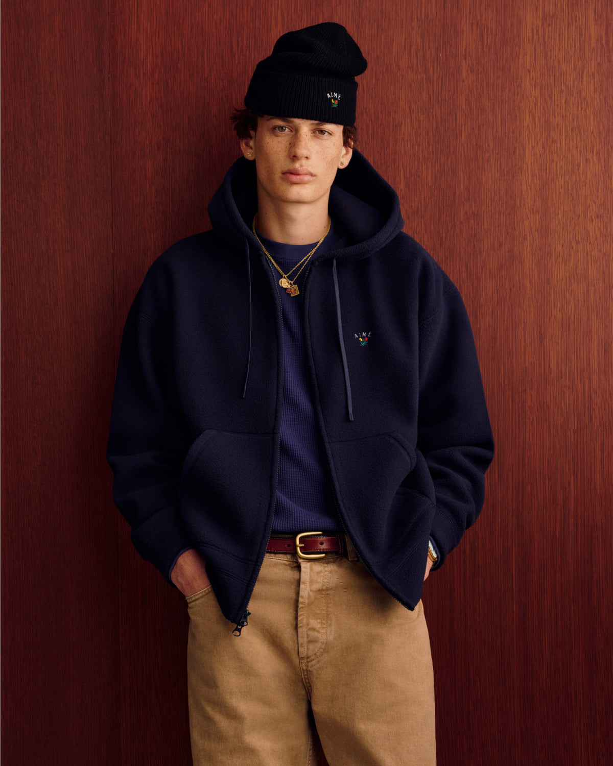 Young person wears Crest Fleece Full-Zip Hoodie, blue shirt, and tan pants against wood-paneled background, hands in pockets.
