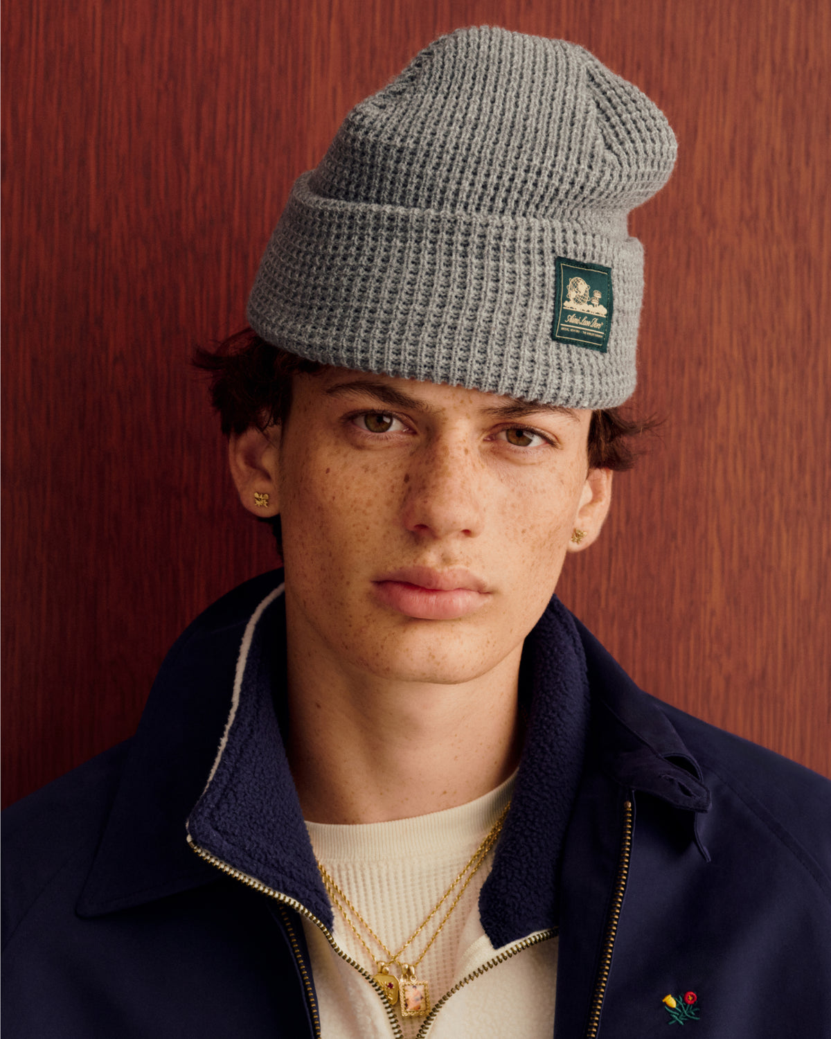 Young man wears a gray Waffle Knit Beanie and navy jacket, standing by wood paneling, looking at the camera.