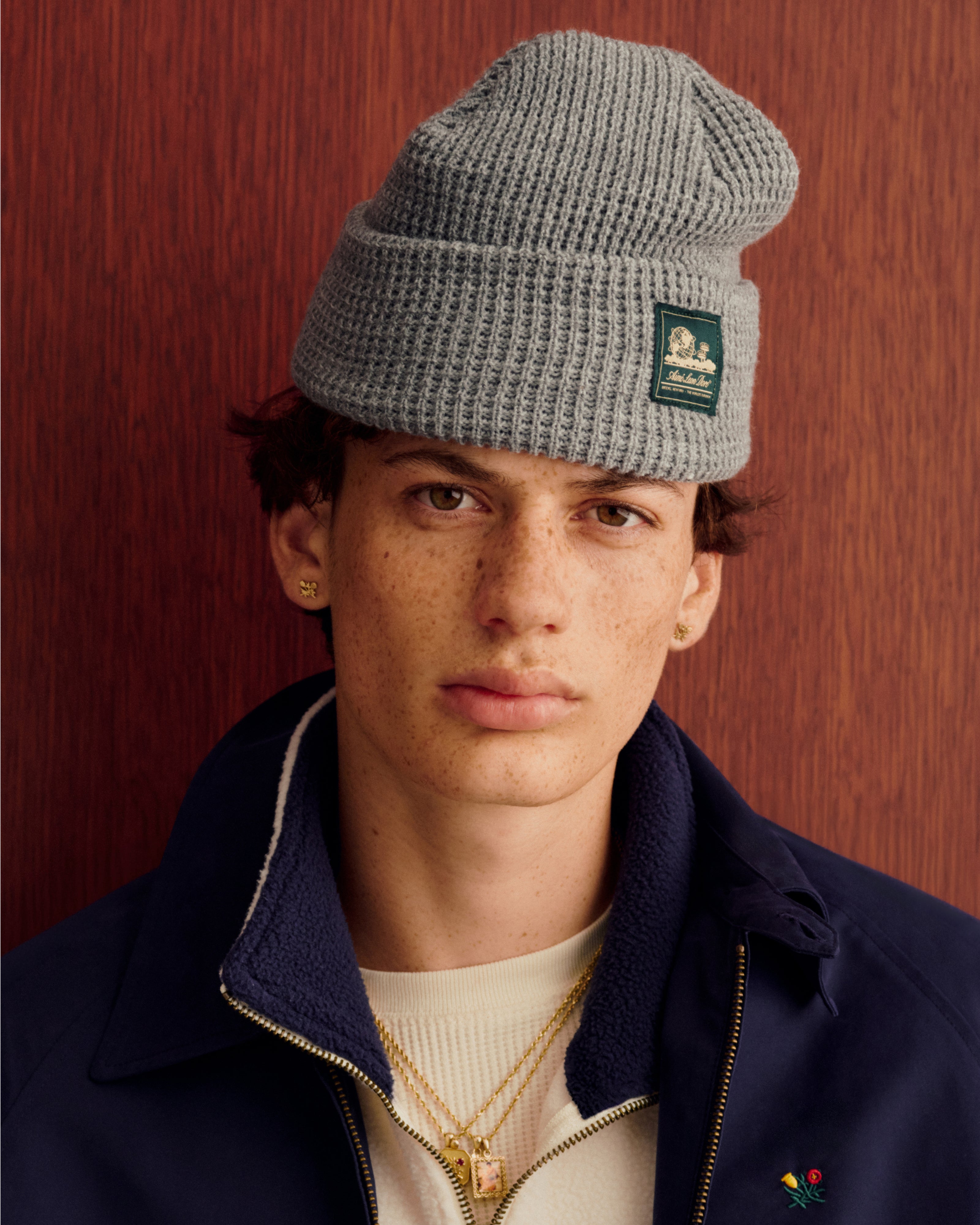 Young man wears a gray Waffle Knit Beanie and navy jacket, standing by wood paneling, looking at the camera.