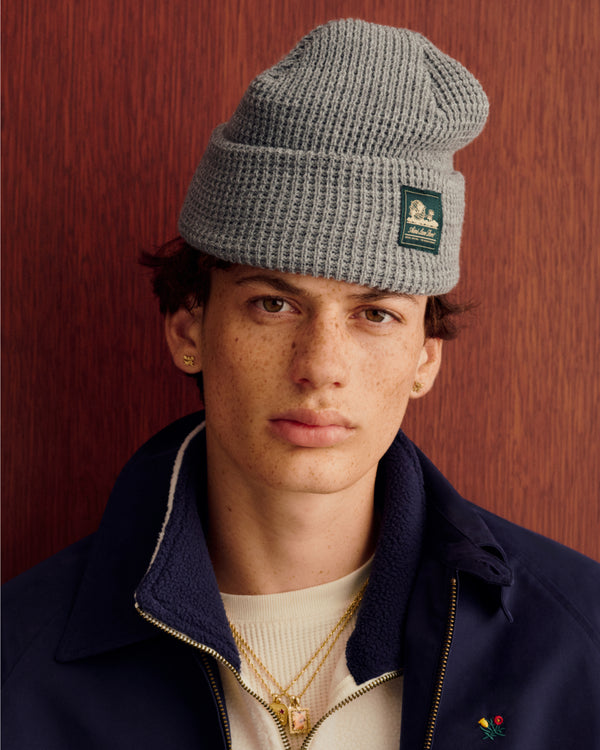 Young man wears a gray Waffle Knit Beanie and navy jacket, standing by wood paneling, looking at the camera.