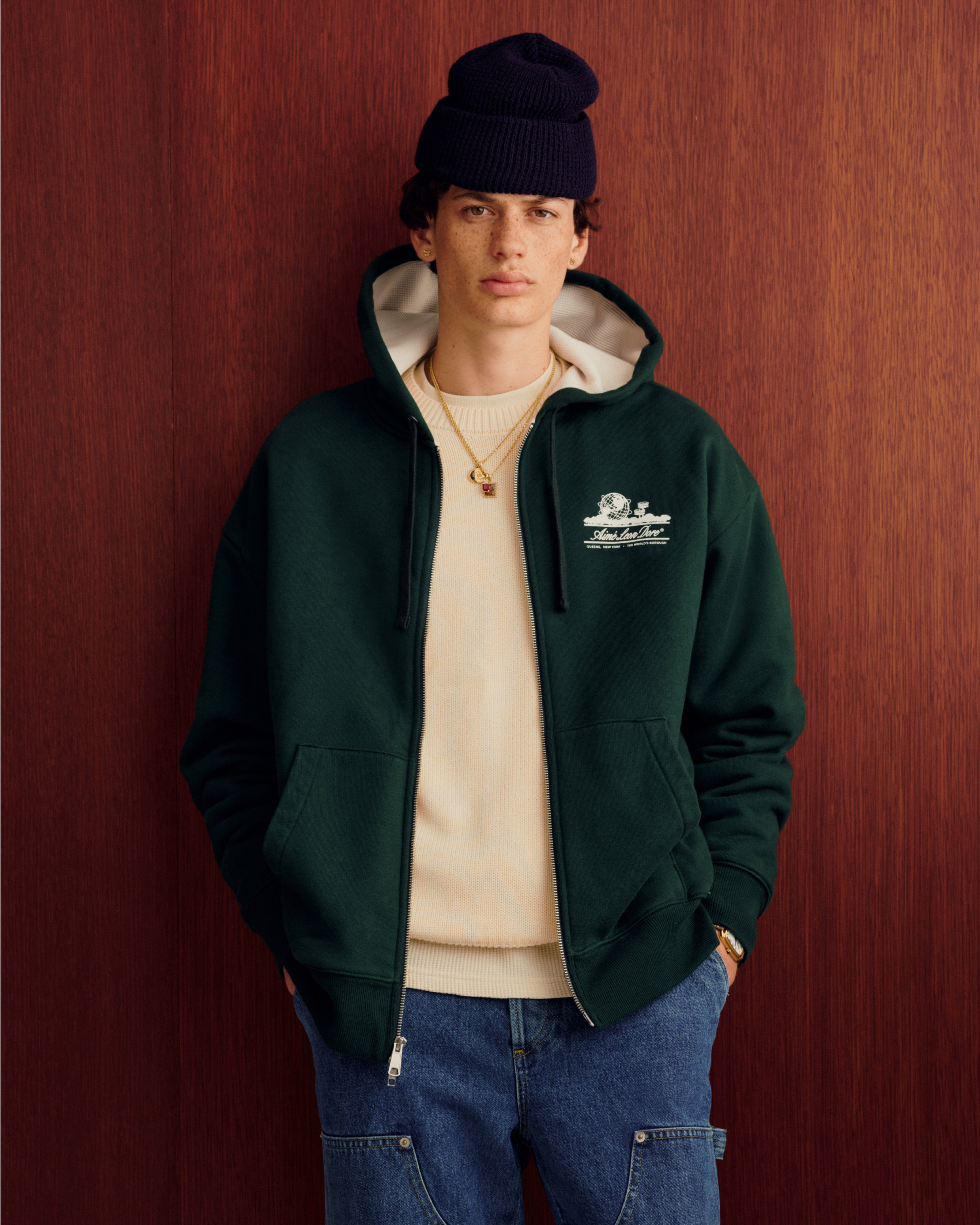 Someone wears the Unisphere Waffle Lined Full-Zip Hoodie, a beige sweater, jeans, and a knit hat by a wood-paneled wall.