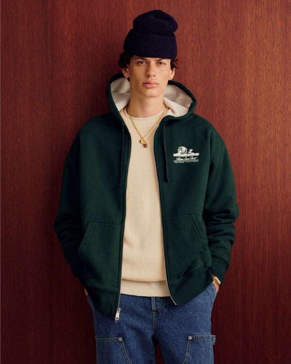Someone wears the Unisphere Waffle Lined Full-Zip Hoodie, a beige sweater, jeans, and a knit hat by a wood-paneled wall.
