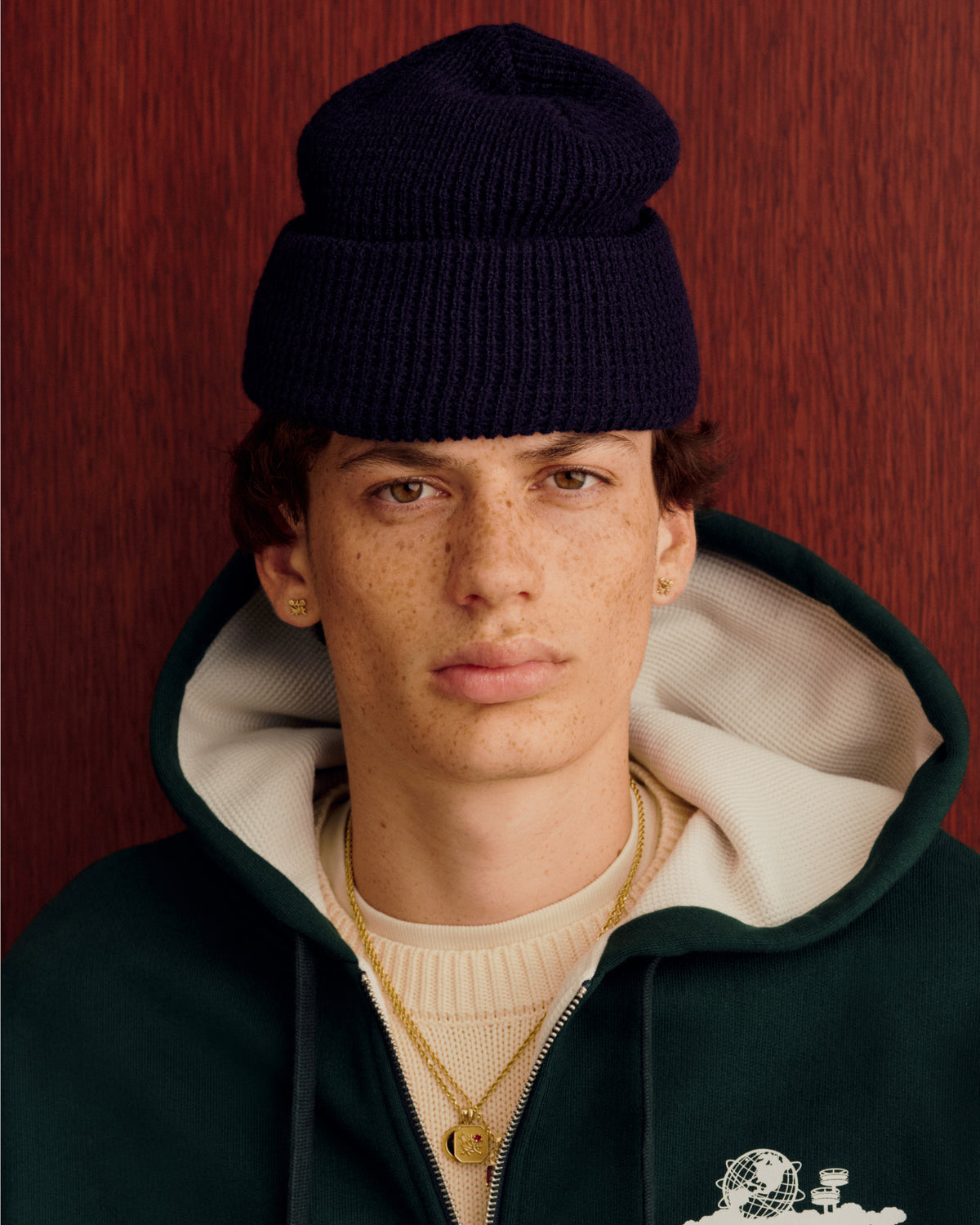Young person with freckles in a Waffle Knit Beanie, green hoodie, layered necklaces, and gold earrings by a red wall.