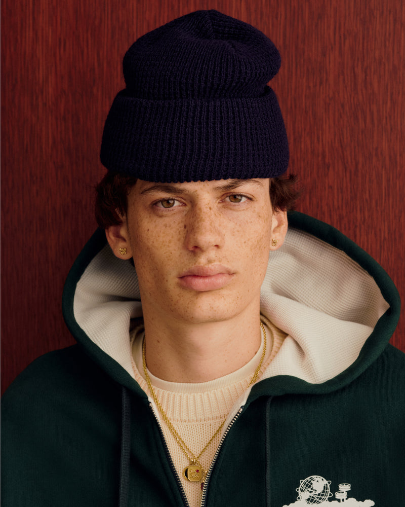 Young person with freckles in a Waffle Knit Beanie, green hoodie, layered necklaces, and gold earrings by a red wall.